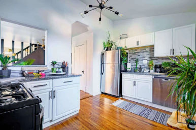 Bright renovated kitchen with white cabinets, stainless-steel fridge and dishwasher, gas stove, gray granite countertops and mosaic tile backsplash, warm hardwood floors, indoor plants, striped runner rug and a black modern chandelier.