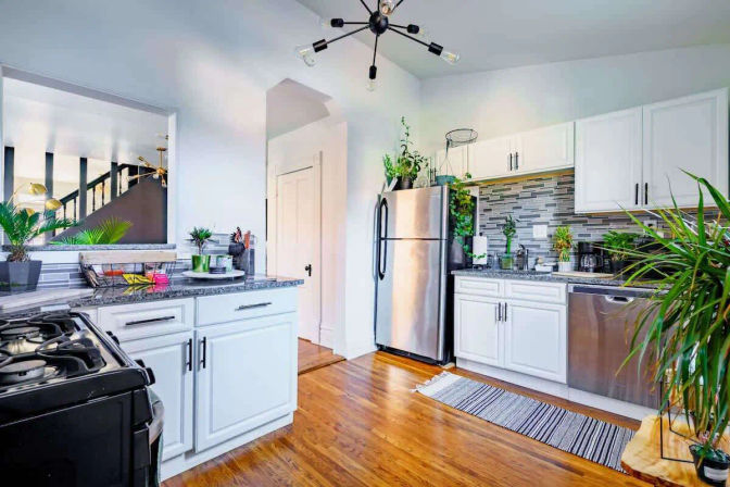 Bright renovated kitchen with white cabinets, stainless-steel fridge and dishwasher, gas stove, gray granite countertops and mosaic tile backsplash, warm hardwood floors, indoor plants, striped runner rug and a black modern chandelier.