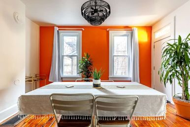 Sunlit urban dining room with bold orange accent wall, two white-trim windows, linen-covered table with metal chairs, potted plants, hardwood floors and a black woven chandelier.
