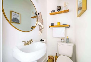 Small modern half-bath with round gold mirror, white pedestal sink and brushed-gold faucet, wooden floating shelves with decor, towel rack above the toilet, and gold-accent accessories.