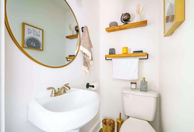 Small modern half-bath with round gold mirror, white pedestal sink and brushed-gold faucet, wooden floating shelves with decor, towel rack above the toilet, and gold-accent accessories.