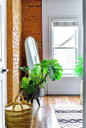 Sunlit cozy apartment nook with exposed brick wall, full-length leaning mirror, large monstera plant in a black planter, woven basket on polished hardwood floors and a patterned rug by a bright white window.