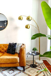 Sunlit modern living room corner with a tan leather sofa, patterned throw pillows, gold arched floor lamp, black side table with potted plant, large leafy houseplant and geometric boho rug.
