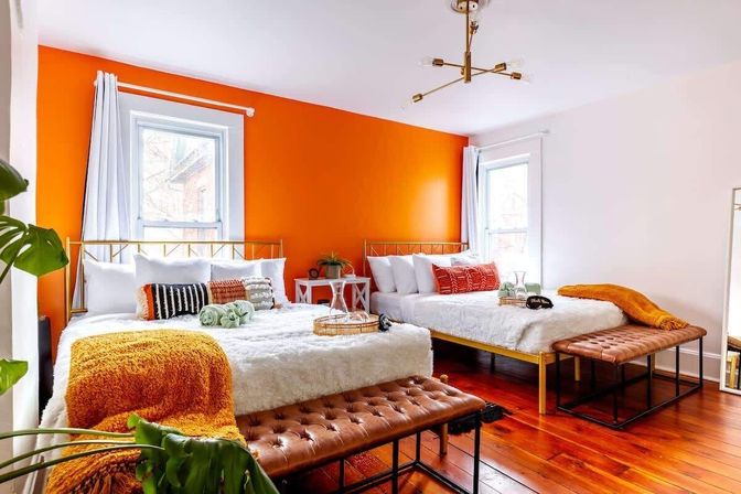 Bright contemporary bedroom interior with a bold orange accent wall, two gold-frame beds dressed in white fluffy bedding, colorful textured pillows and mustard throws, tufted leather benches, warm hardwood floors, brass chandelier and a potted plant — cozy guest room with a pop of orange.