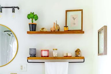 Two wooden floating shelves on a bright white bathroom wall above a black towel bar with a white towel, featuring a potted plant, framed cocktail print, candles, a mason jar of cotton swabs, small ceramics and figurines, with a round gold mirror at left.