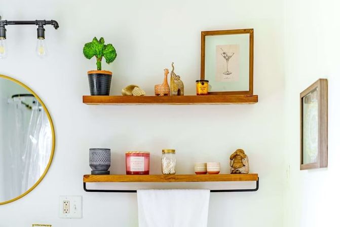 Two wooden floating shelves on a bright white bathroom wall above a black towel bar with a white towel, featuring a potted plant, framed cocktail print, candles, a mason jar of cotton swabs, small ceramics and figurines, with a round gold mirror at left.