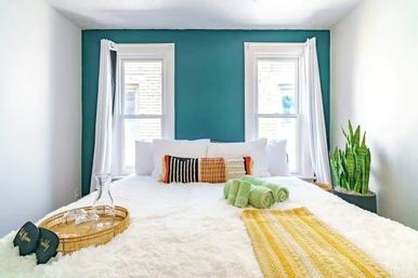 Sunlit modern bedroom with teal accent wall and two windows, plush white bed layered with woven accent pillows, yellow runner, rolled green towels, wicker tray with glass carafe, and a snake plant by the window.