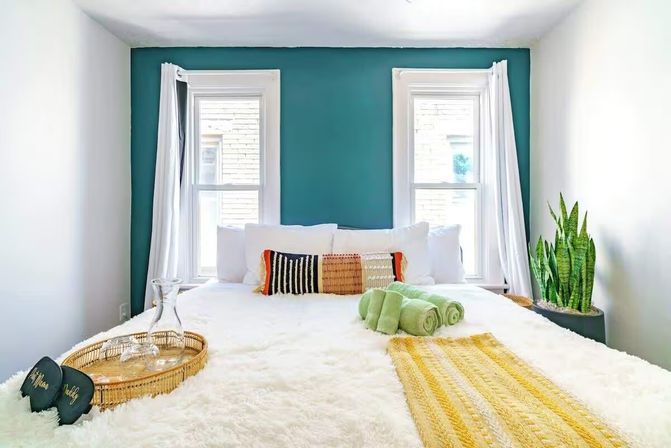 Sunlit modern bedroom with teal accent wall and two windows, plush white bed layered with woven accent pillows, yellow runner, rolled green towels, wicker tray with glass carafe, and a snake plant by the window.