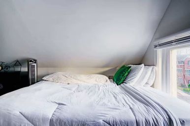 Cozy attic loft bedroom with sloped ceiling, bright white bedding, a green accent pillow, tower fan on a nightstand, and a large window revealing nearby brick apartment buildings.