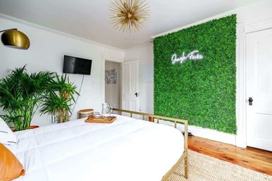 Bright modern bedroom with white bedding and gold metal bed frame, lush green living wall with neon sign, potted palms and hardwood floor