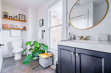 Sunlit modern bathroom with black vanity and brass faucet, round gold mirror, white toilet, wood floating shelves, gray tile floor and a large green monstera plant by the window.