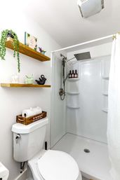 Compact white bathroom with corner walk-in shower and square rainfall showerhead, wooden floating shelves with a trailing plant, and a toilet topped with a wooden crate of toilet paper.
