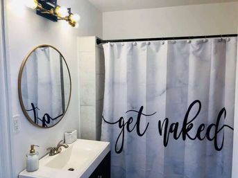 Bright modern bathroom with round brass-framed mirror, white vanity and soap dispenser, and a gray marble-look shower curtain on a black rod printed with playful black script saying 'get naked'.