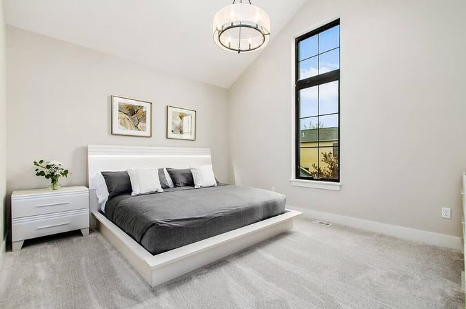 Bright modern minimalist bedroom with a white low platform bed dressed in gray bedding, white nightstand with vase of flowers, tall black-framed window overlooking the neighborhood, and round pendant light