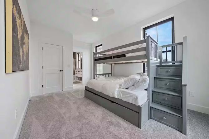 Bright modern bedroom with gray twin-over-full bunk bed, stair-style drawers and trundle, plush light-gray carpet, white walls, large windows and ceiling fan — family-friendly kids’ room.