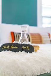 Black sleep masks with gold script reading "Plant Mama" and "Daddy" resting on a fluffy white bedspread, with a clear glass carafe and colorful decorative pillows blurred in the cozy bedroom background.