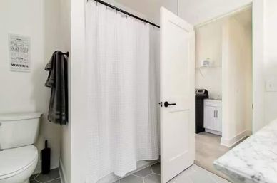 Bright modern bathroom with white shower curtain, hex gray floor tiles, toilet and dark gray towels; open door shows adjacent laundry area with washing machine and white cabinets.
