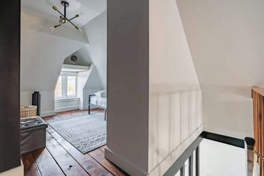 Cozy upstairs attic-style bedroom with sloped ceilings and dormer window, white walls, modern sputnik chandelier, gray patterned rug over wide dark wood plank floors, simple metal-frame bed and nearby stair railing.