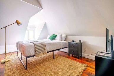 Cozy attic bedroom with sloped ceiling, metal-frame bed dressed in white linens, green pillow and striped throw, braided jute rug on hardwood floors, brass floor lamp, black nightstand and TV.