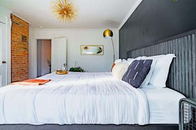 Modern urban loft bedroom with king-size white bedding, exposed brick column, dark gray accent wall, gold sputnik chandelier, floor lamp, and decorative pillows.