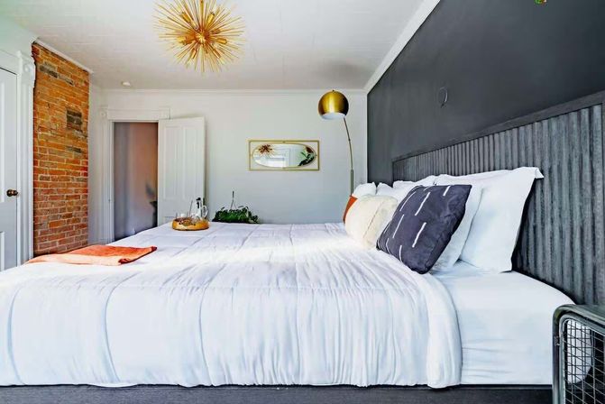Modern urban loft bedroom with king-size white bedding, exposed brick column, dark gray accent wall, gold sputnik chandelier, floor lamp, and decorative pillows.