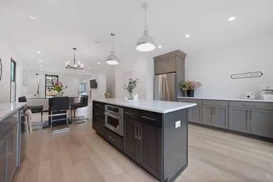 Bright open-concept modern kitchen with large dark island, white quartz countertops, stainless steel refrigerator, gray cabinetry, pendant lights, hardwood floors and a coffee bar sign.