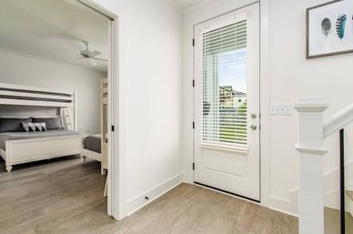 Bright modern entryway with glass-paneled door and blinds, white walls, light wood floors, staircase, and an adjacent bedroom with a white bunk bed and ceiling fan — minimalist coastal farmhouse style.