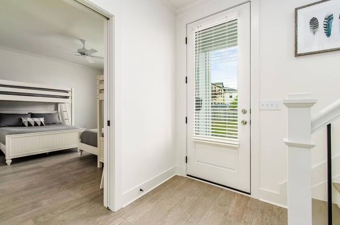 Bright modern entryway with glass-paneled door and blinds, white walls, light wood floors, staircase, and an adjacent bedroom with a white bunk bed and ceiling fan — minimalist coastal farmhouse style.