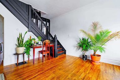 Bright home interior with honey-toned hardwood floors, black-painted staircase, small red desk and chair tucked under the stairs, and several potted green plants creating a cozy home office nook.