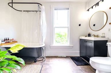 Bright modern farmhouse bathroom with black clawfoot tub and clear shower curtain, black vanity topped with round gold mirror, large central window showing greenery, indoor plant and wood-look tile floor