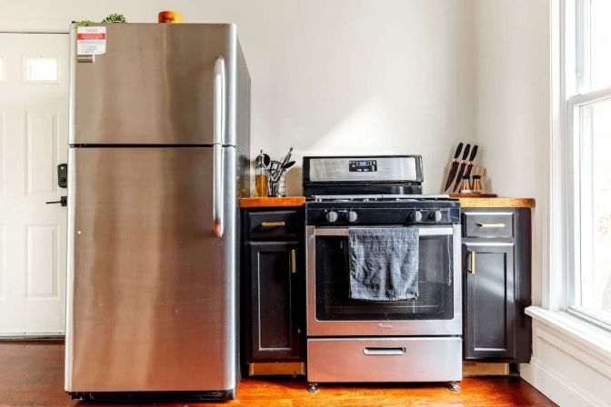 Sunlit modern kitchen with stainless steel top-freezer refrigerator beside a stainless gas range draped with a dark towel, black cabinets, butcher-block countertops and hardwood floor by a large window.