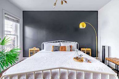 Bright modern urban bedroom with white metal bed, textured white bedding, charcoal accent wall, rattan nightstands, gold arc floor lamp and potted palm by the window.