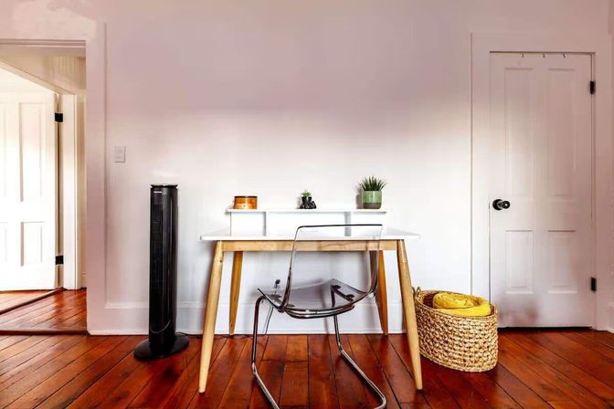 Minimalist home office nook with white desk, clear chair, potted succulents, woven basket and tower fan on warm hardwood floors and white walls.