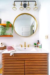 Bright modern bathroom vanity with white countertop, gold faucet and round gold-framed mirror, slatted wood cabinet, glass globe light fixture, stacked green towels on an open wood shelf and a small potted succulent.
