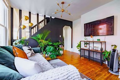 Sunlit modern living room with green velvet sofa, cozy throws and pillows, abundant indoor plants, black accent staircase wall, gold mid-century chandelier and floor lamp, wall-mounted TV and warm hardwood floors.