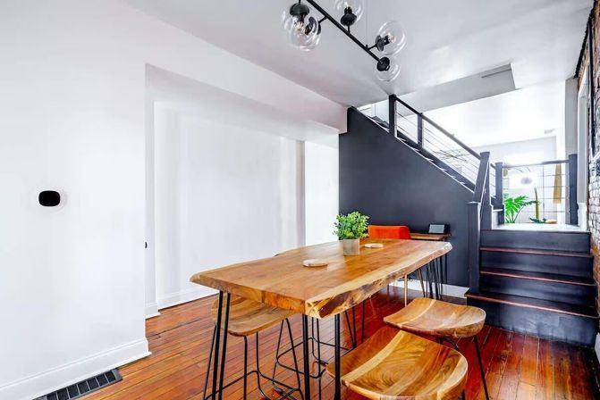 Bright modern loft dining area with live-edge wooden table and matching curved wood stools, black staircase wall, glass globe pendant light, hardwood floors and small potted plant centerpiece