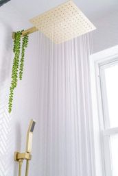 Gold square rainfall showerhead pouring water in a modern white-tiled spa-like bathroom with matching gold handheld shower, hanging green plant, and window light.