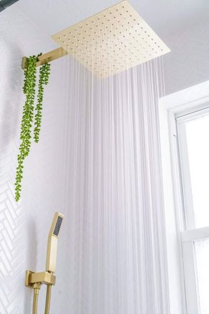 Gold square rainfall showerhead pouring water in a modern white-tiled spa-like bathroom with matching gold handheld shower, hanging green plant, and window light.