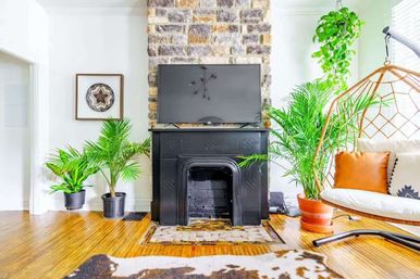 Sunny boho living room with a stone hearth and black fireplace mantel topped by a flat-screen TV, lush potted palms, rattan hanging chair with an orange pillow, and warm hardwood floors.
