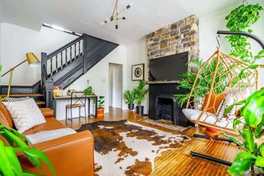 Bright modern boho living room with tan leather sofa, cowhide rug, stone fireplace with wall-mounted TV, hanging rattan swing chair, black-painted staircase, hardwood floors and abundant indoor plants
