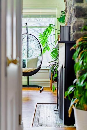 View through an open doorway into a sunlit cozy nook with a black hanging egg chair on a stand, striped cushion, large leafy houseplants, a stone fireplace and warm hardwood floors.