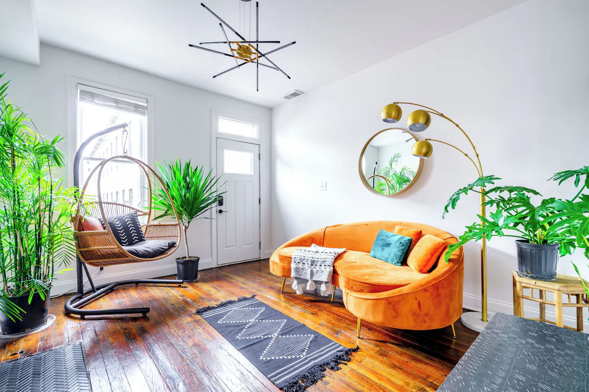 Sunny modern boho living room and entryway with an orange velvet curved sofa, teal accent pillow, rattan hanging chair, abundant indoor plants, round wall mirror, gold arc floor lamp, dark hardwood floors and geometric rug.