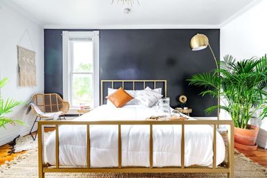 Cozy modern boho bedroom interior with a gold metal bed frame and white bedding against a black accent wall, rattan chair and woven wall hanging, large potted palms, brass floor lamp and sunlit window.