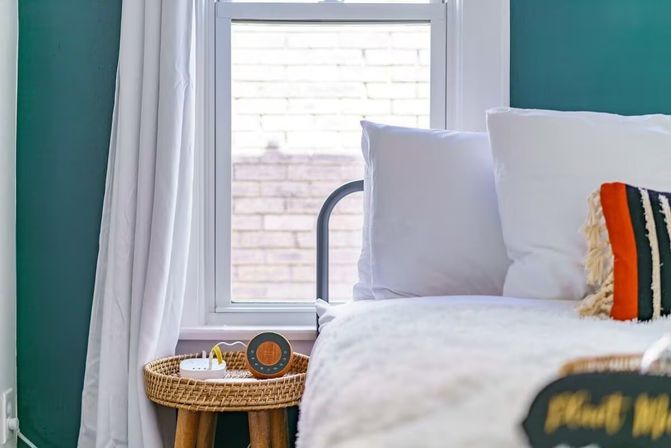 Cozy modern bedroom corner with teal accent wall, white curtains and fluffy bedding, wicker bedside stool holding a round portable speaker and small charger, and a colorful fringed throw pillow by a window looking out to a brick exterior.