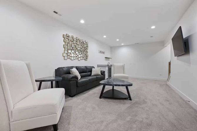 Bright finished basement living area with gray carpet, black leather sofa, two white accent chairs, round black coffee table, wall-mounted TV, mirrored geometric wall art and small wet bar