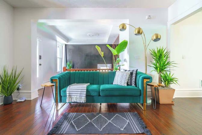 Sunlit modern living room with a teal velvet sofa, patterned throw and pillows, gold arc floor lamp, large potted houseplants, dark hardwood floors and a geometric rug