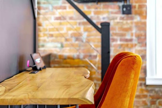 Cozy loft-style workspace with a live-edge wooden desk, orange velvet chair, exposed brick wall and industrial lamp