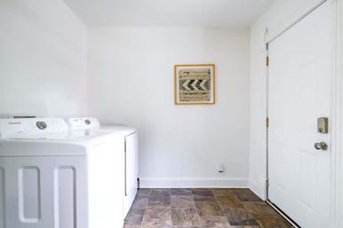 Bright minimal residential laundry room with white top-load washer and dryer, framed woven wall art, brown tile flooring and a white entry door.