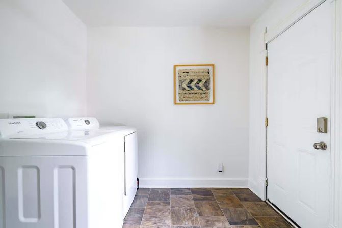 Bright minimal residential laundry room with white top-load washer and dryer, framed woven wall art, brown tile flooring and a white entry door.