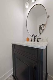 Sleek small powder room with a black vanity topped in white marble, matte black faucet, amber soap dispenser and large round wall mirror under pendant lights.